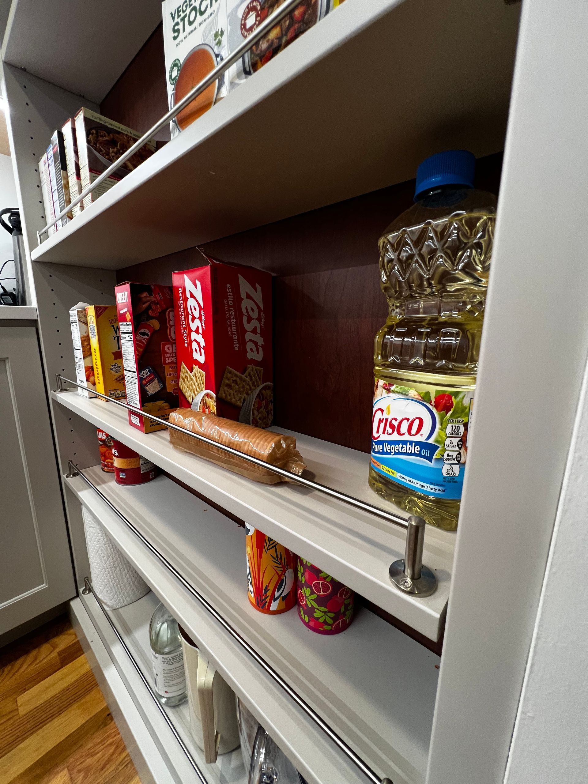 Pantry shelving featuring food items like Zesta crackers, a bottle of Crisco oil, and canned goods behind a metal rail.
