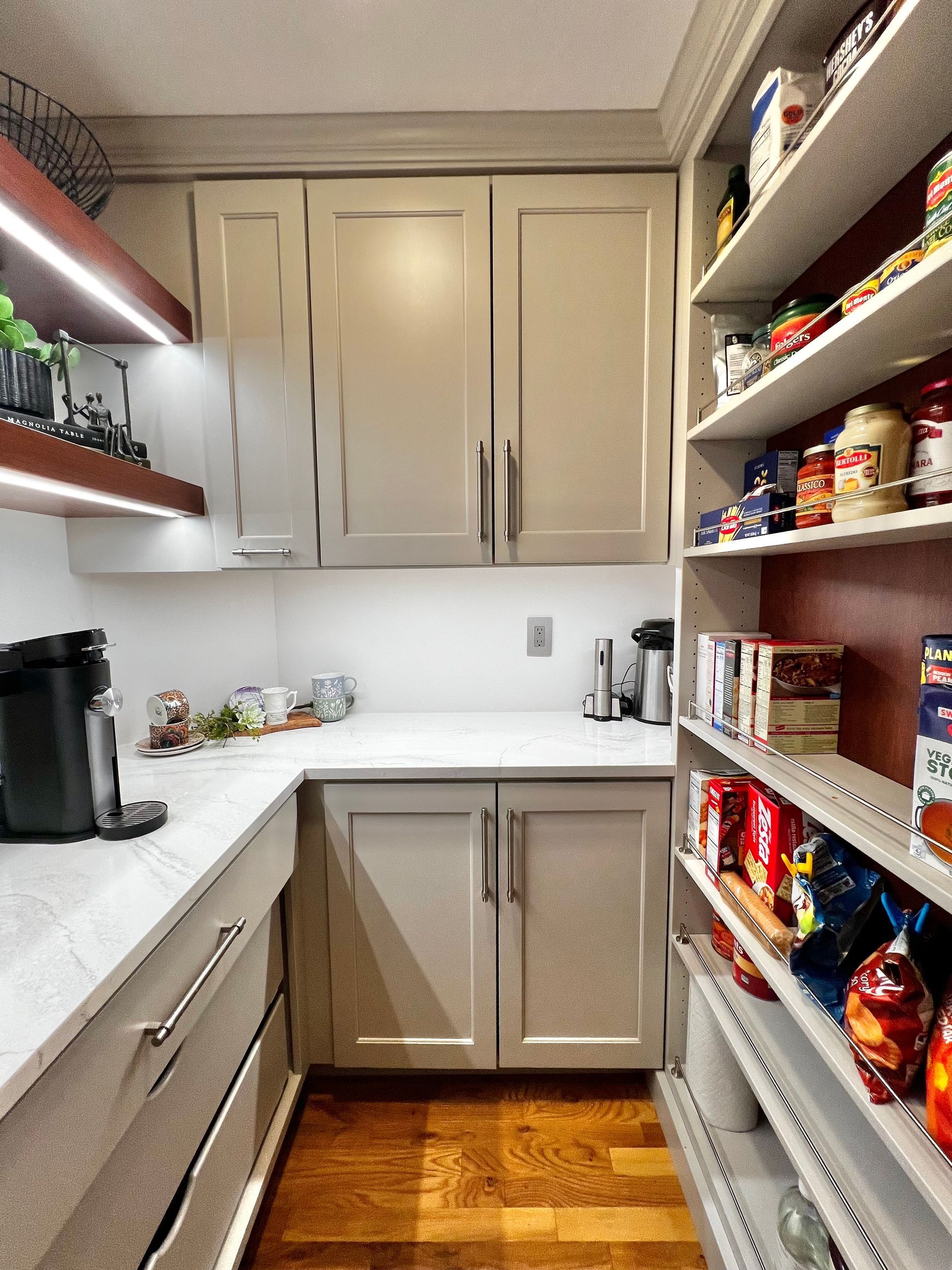 A kitchen pantry with white countertops, light gray cabinets, and open shelving filled with food items on the right.
