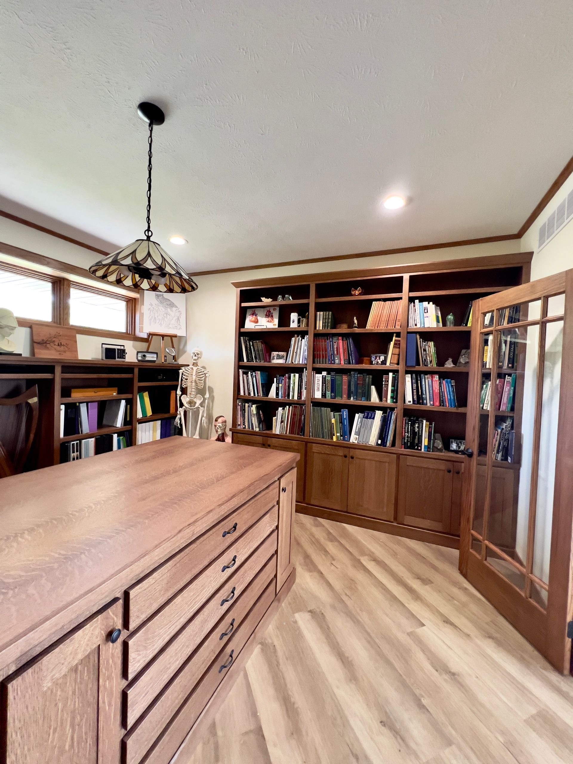 Home office with wooden bookshelves, a large wooden desk with drawers, wood-look flooring, and a stained-glass pendant.