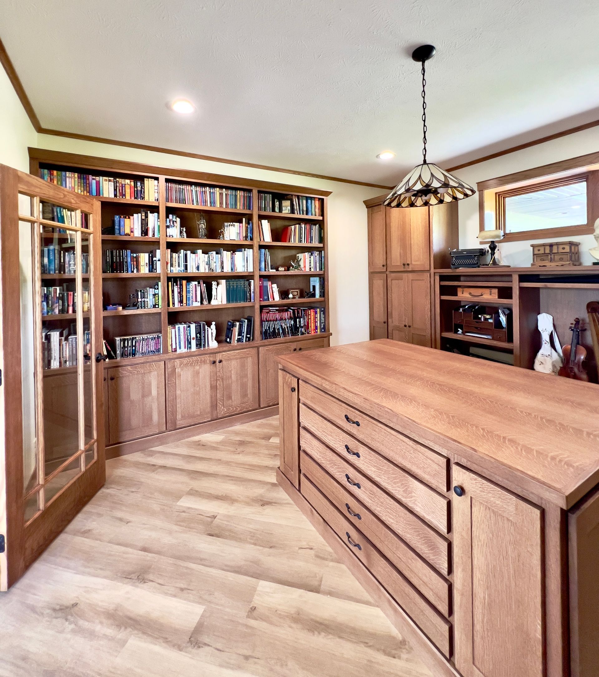 A home office with light wood floors, a large wooden desk with drawers, and built-in bookshelves filled with books.