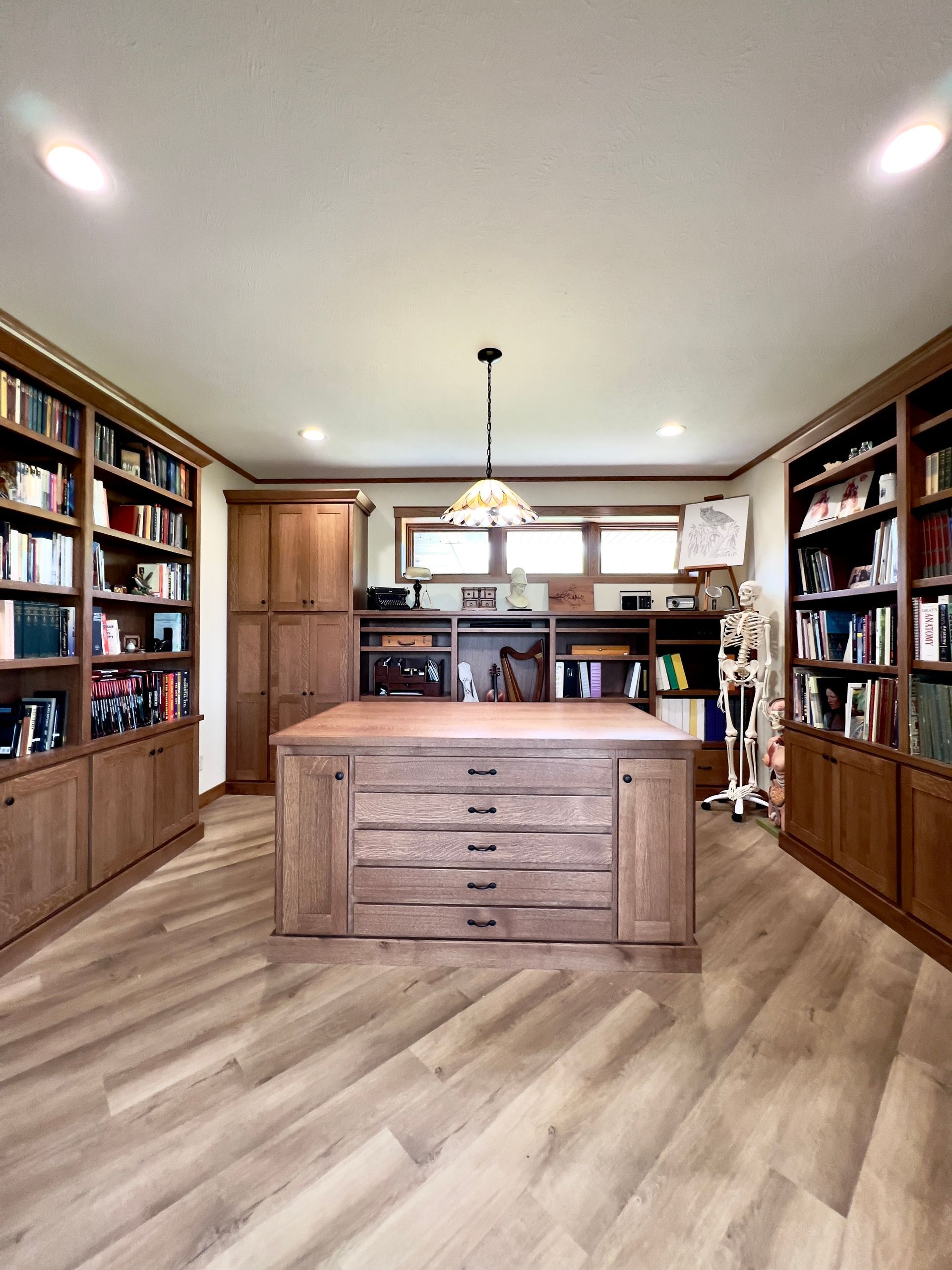 A home office features wood shelving filled with books, a large central wooden desk with drawers, and a wooden skeleton.