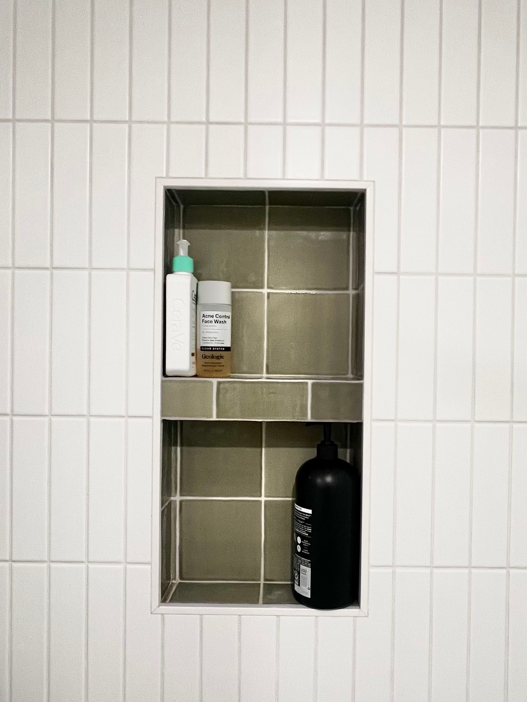 A built-in shower niche with sage green tiles, featuring two shelves holding toiletry bottles against a white tiled wall.