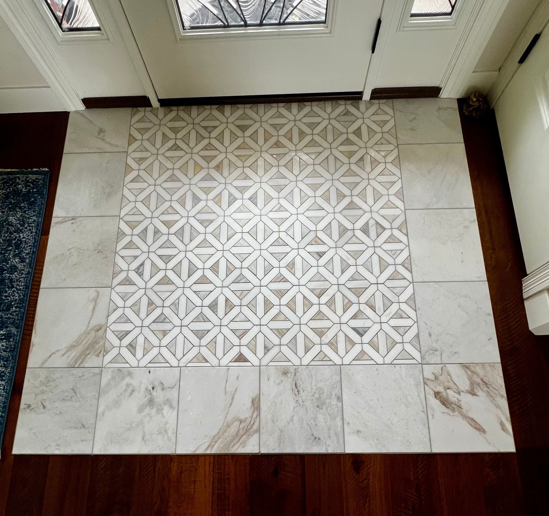 A foyer floor featuring a central square of geometric patterned tiles surrounded by white marble tiles against wood floors.