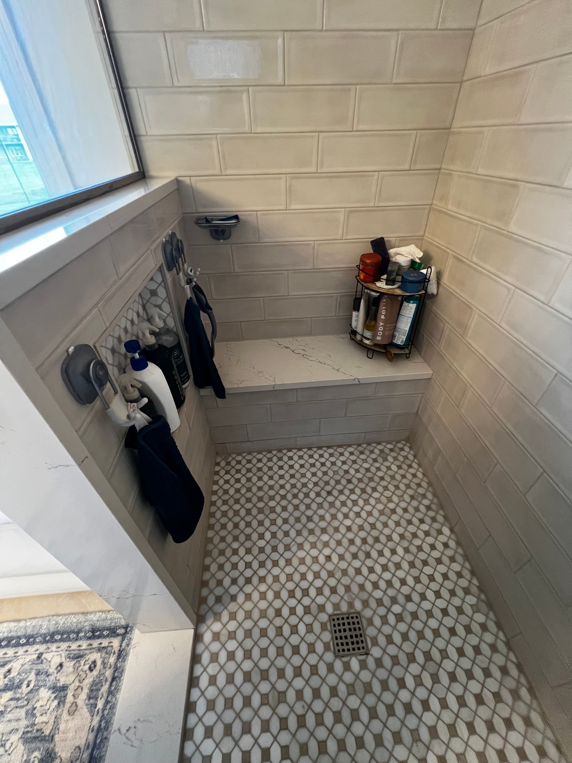 A tiled walk-in shower with a built-in stone bench, patterned floor tiles, and various toiletries stored on the seat.