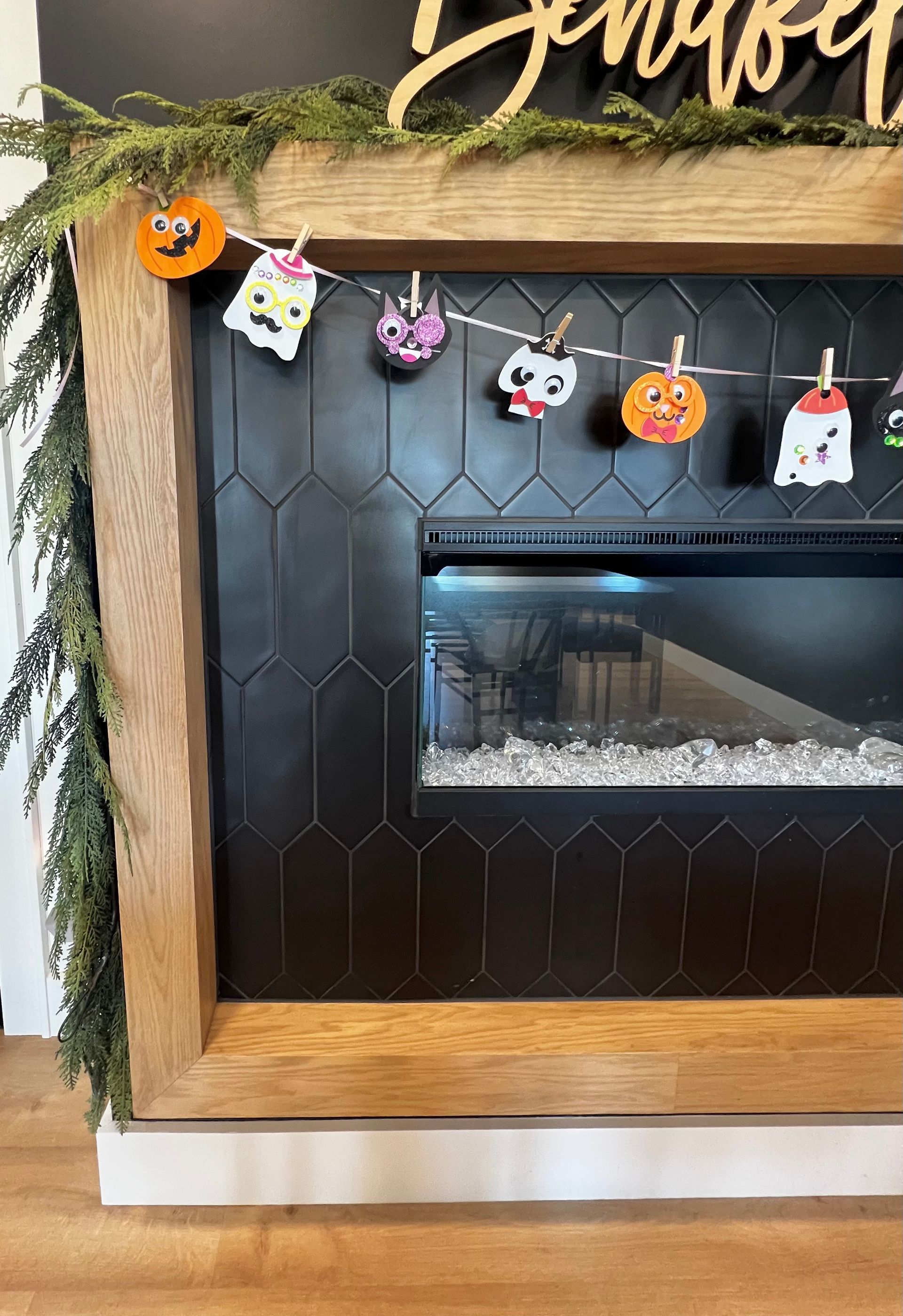 A wooden fireplace mantle with black hexagonal tile, draped with greenery and a festive Halloween garland of icons.