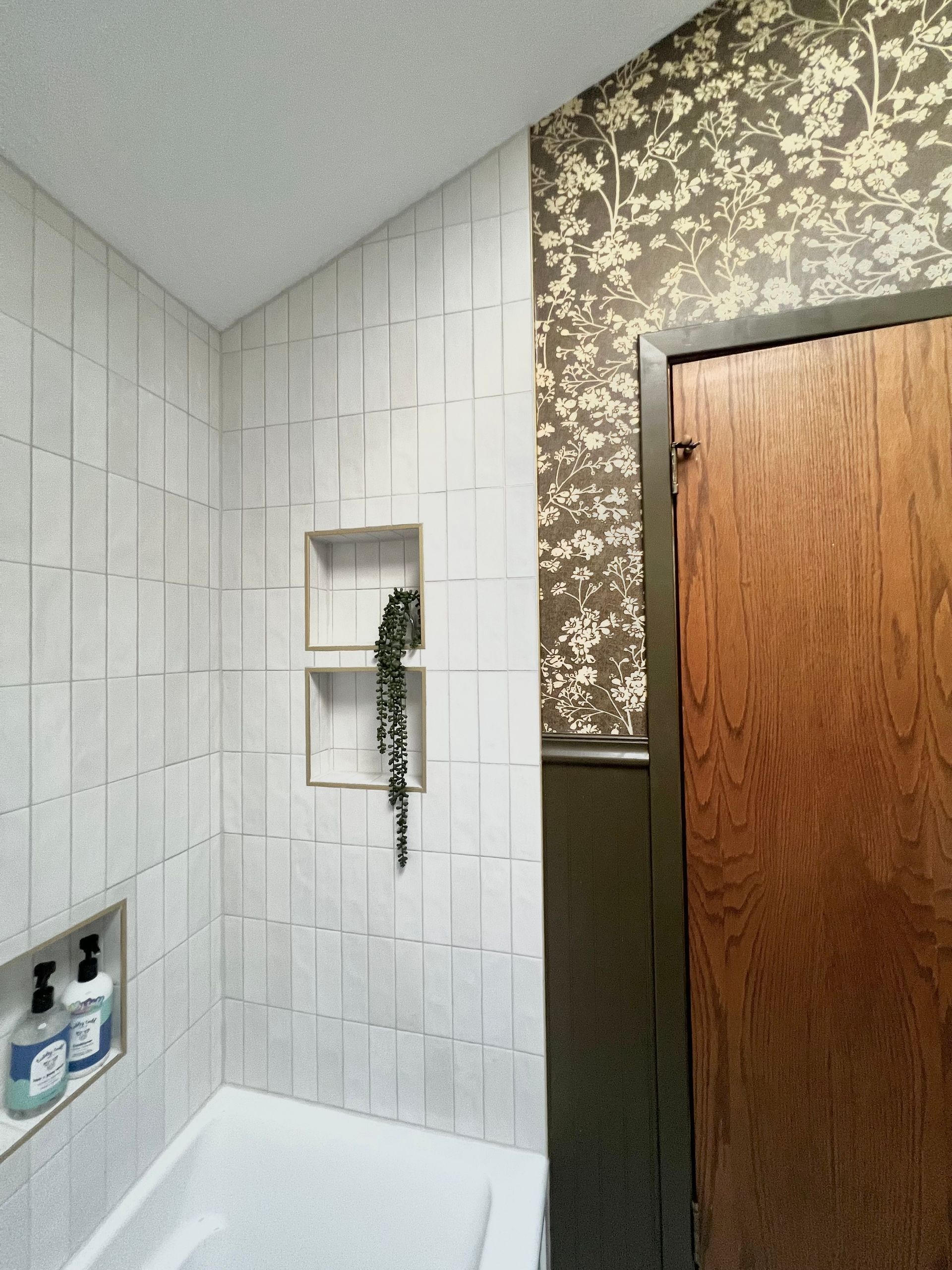 A bathtub shower area with vertical white tiles, built-in wall niches, and an adjacent wall with dark floral wallpaper.