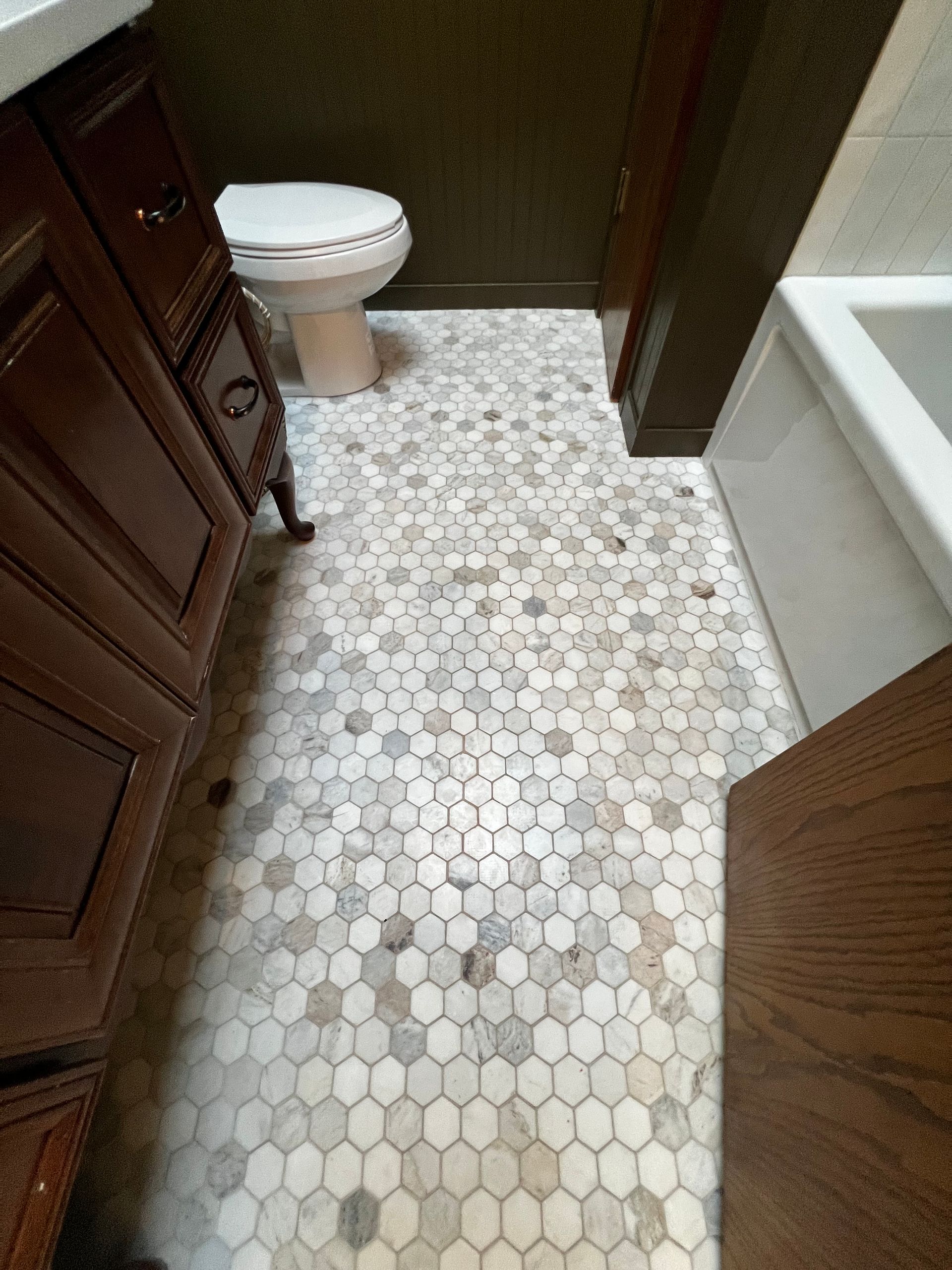 A bathroom floor with light-colored hexagonal stone tiles, flanked by a dark wood vanity on the left and a tub on the right.