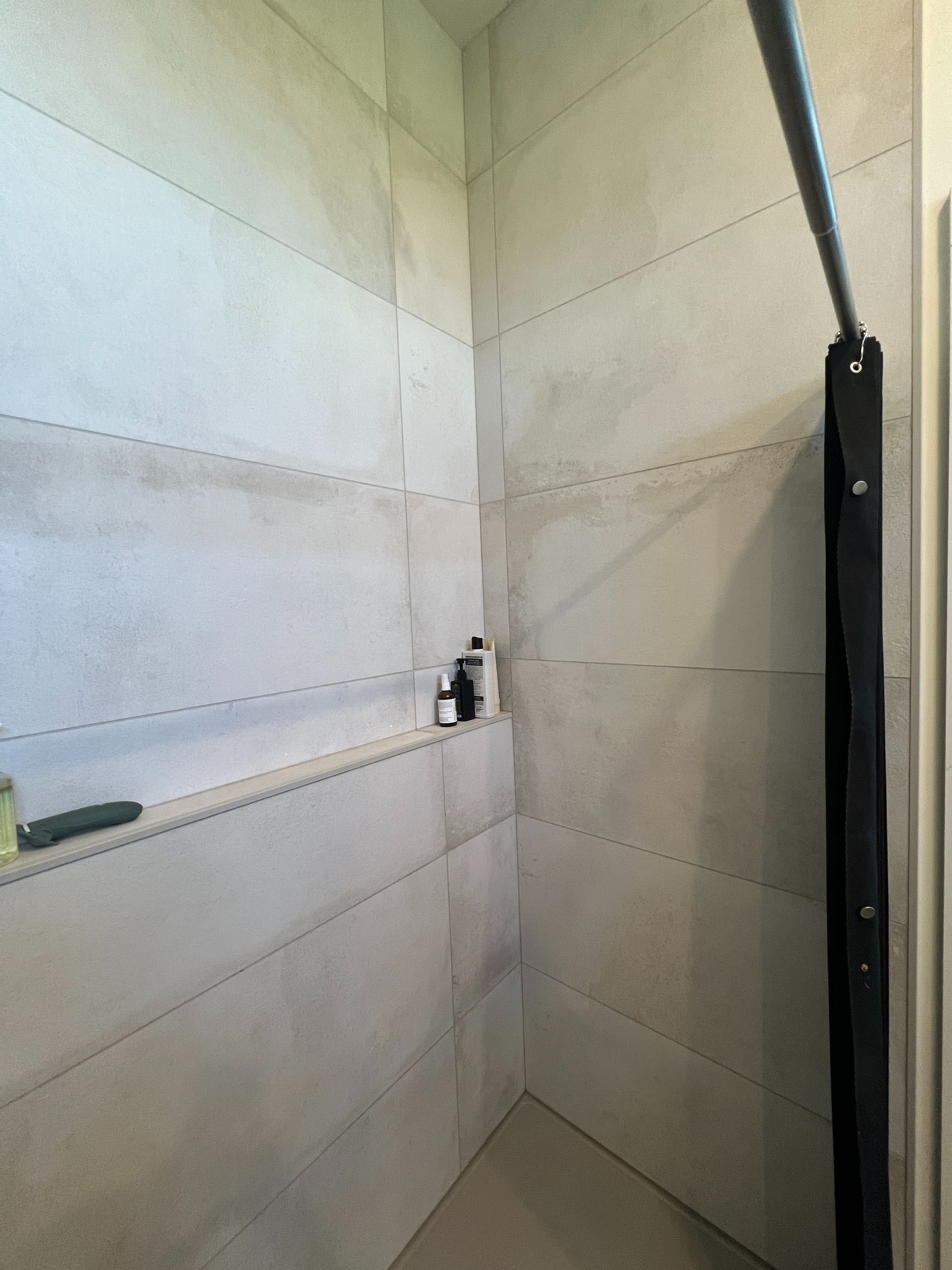 A modern shower with large, light gray tiled walls, a built-in recessed shelf for toiletries, and a dark shower curtain.