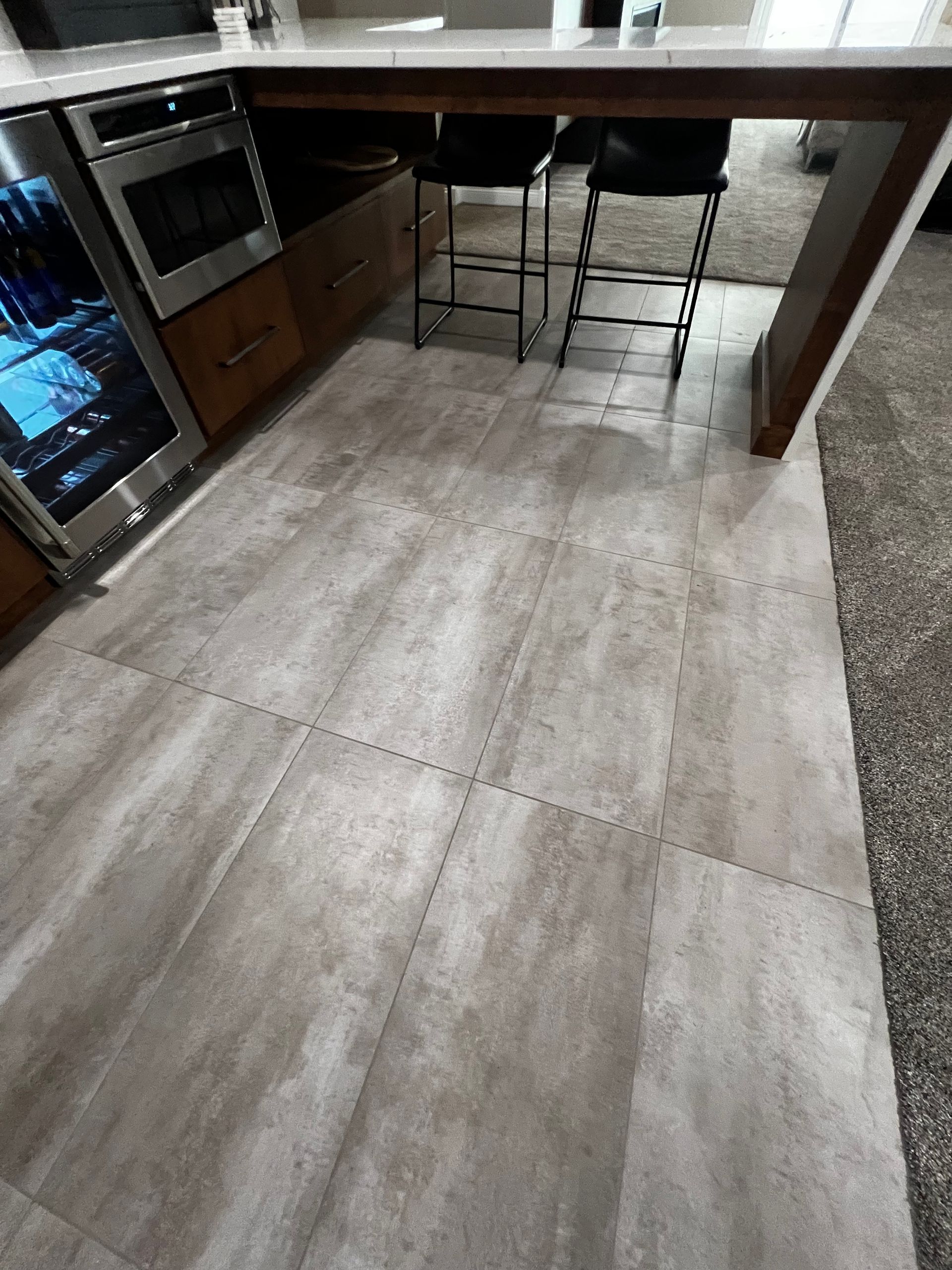A kitchen area featuring grey-toned stone floor tiles, a built-in oven, a wine cooler, and two bar stools under a counter.