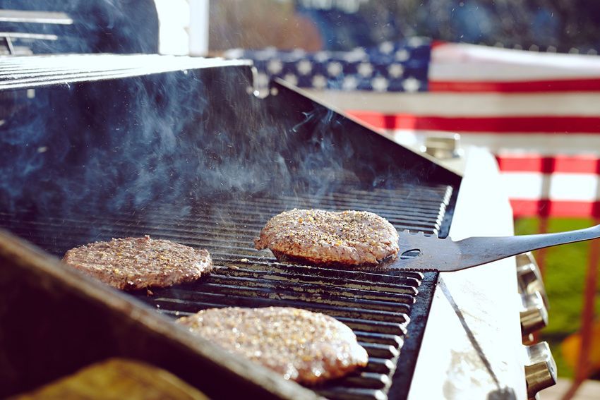 Burgers cooking on a grill with American flag in the background. Smoke rising.