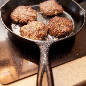 Four hamburger patties are cooking in a cast iron skillet on a stove.