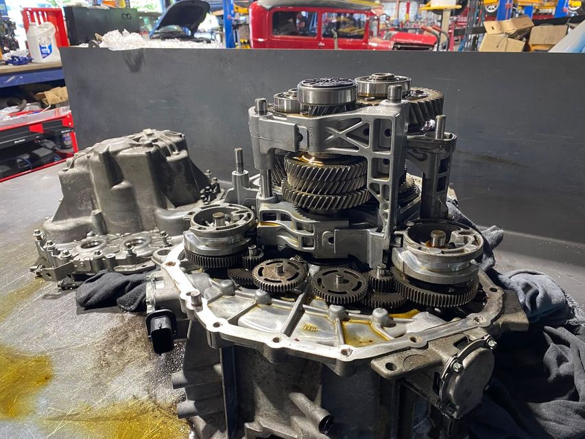 Disassembled automatic transmission on a workbench in a repair shop, with gears and metal parts exposed.