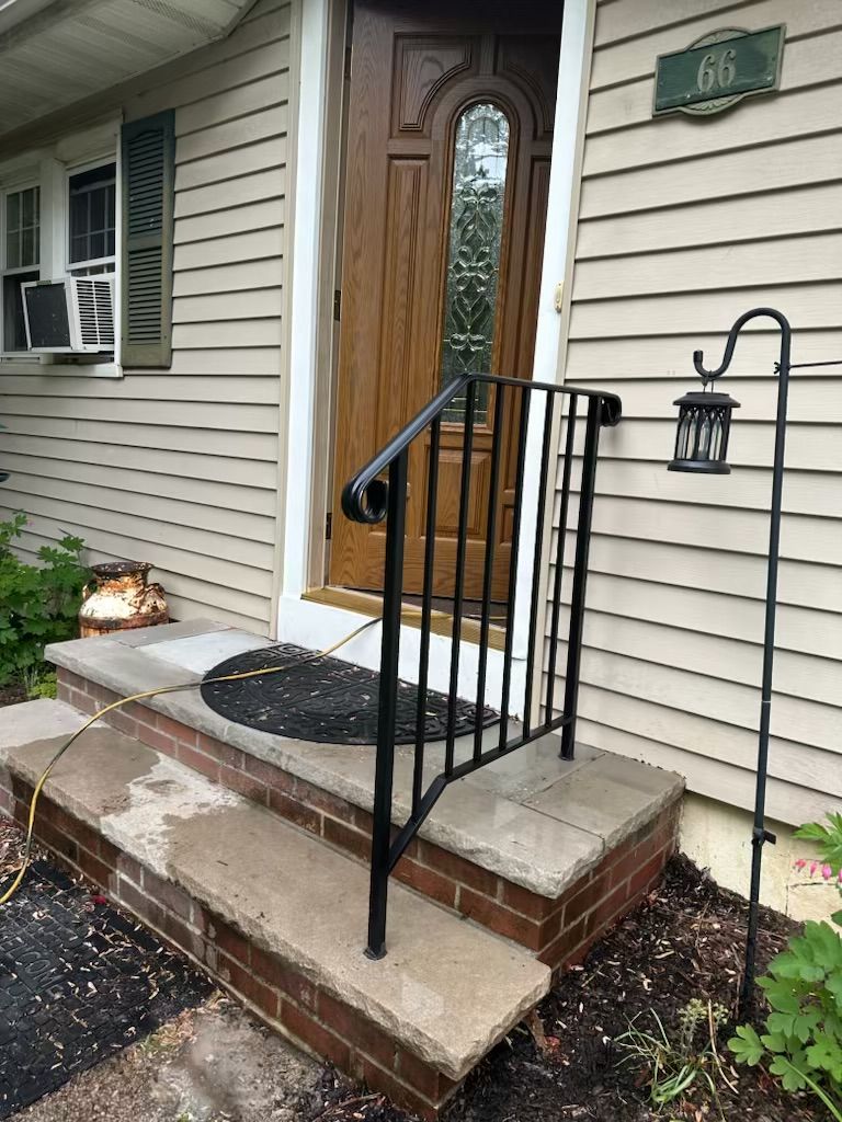Front porch with wooden front door, concrete steps, and black exterior stair safety railing for mobility access