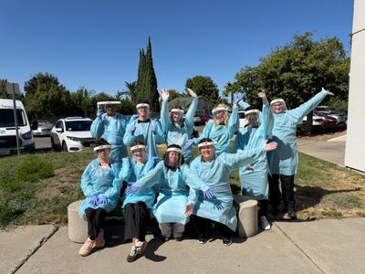 Group of people in light blue shirts posing outdoors on a sunny day, smiling with arms raised