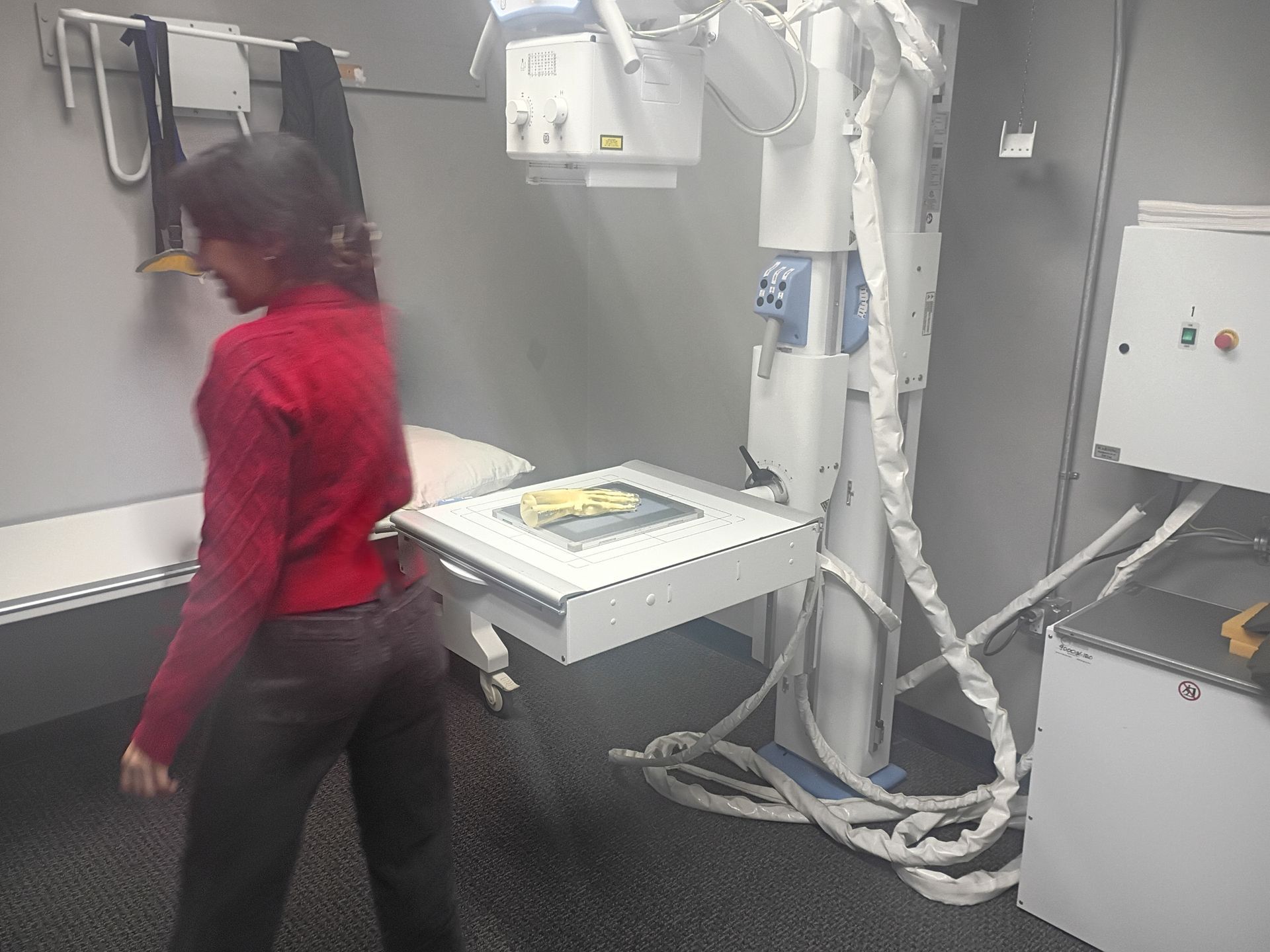 Woman in a red sweater stands in a medical examination room near X-ray equipment.