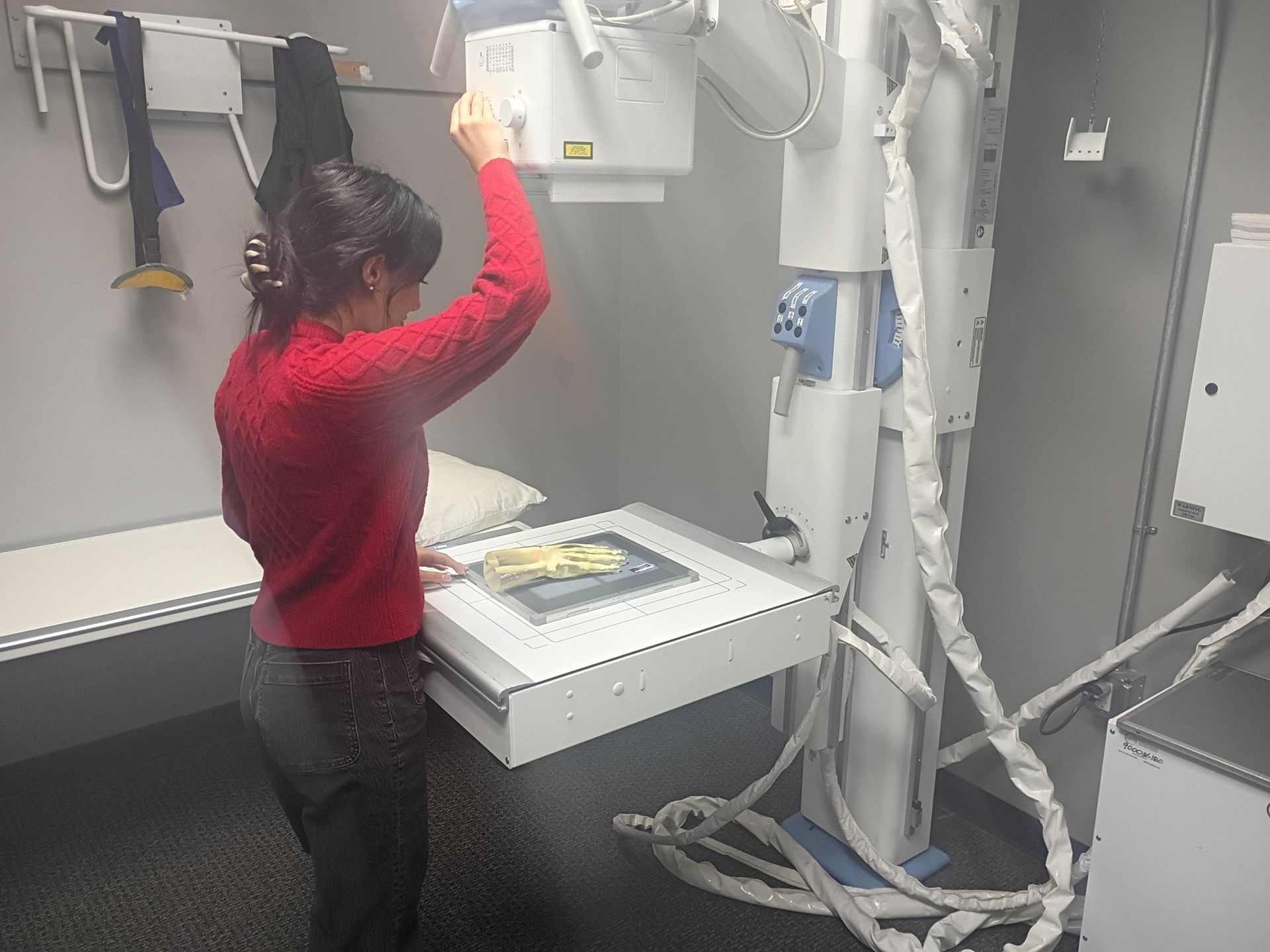 Woman in red shirt adjusts an X-ray machine over a patient table in a medical room.