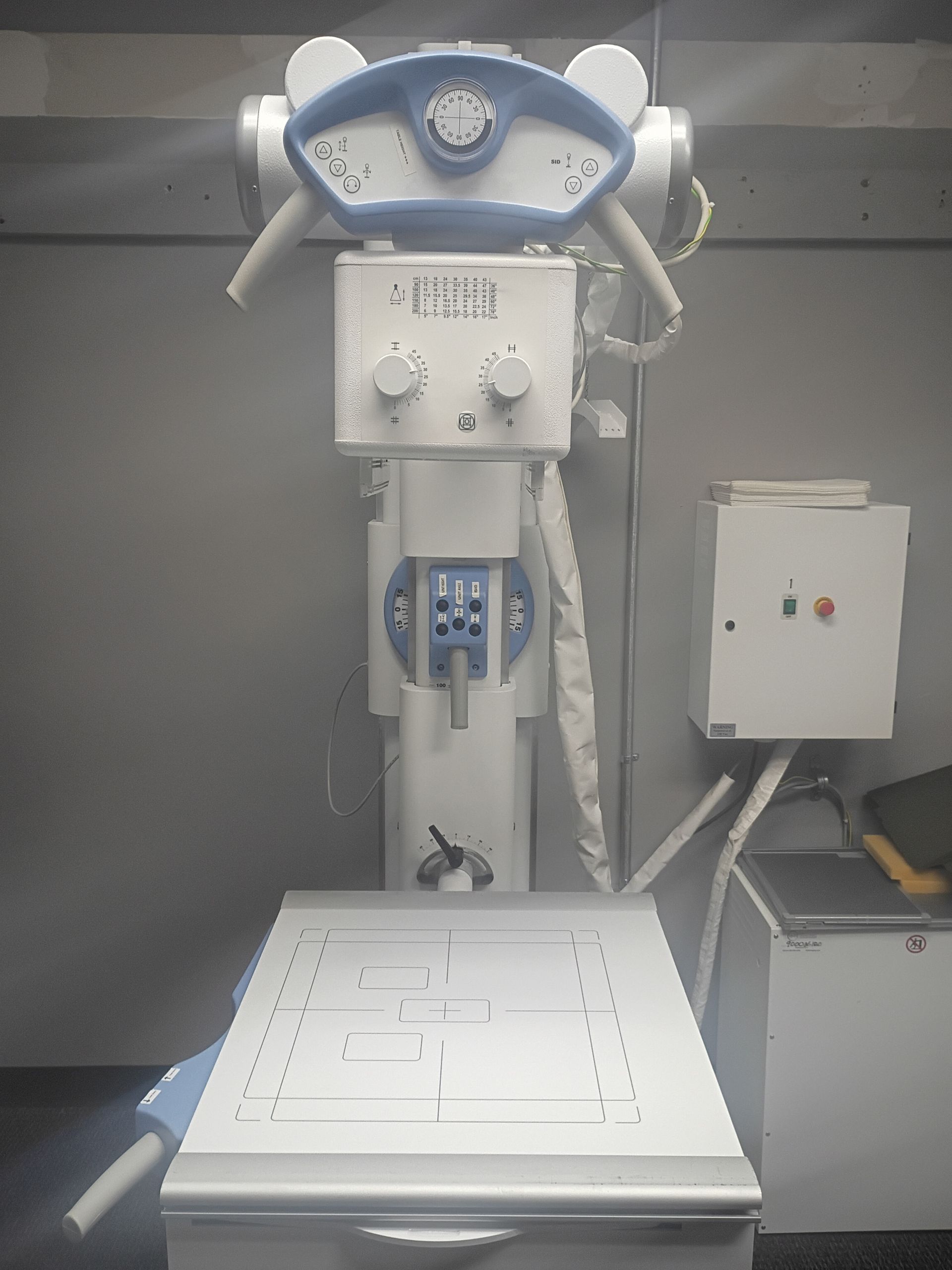 An X-ray machine with a white table and controls, against a gray wall.