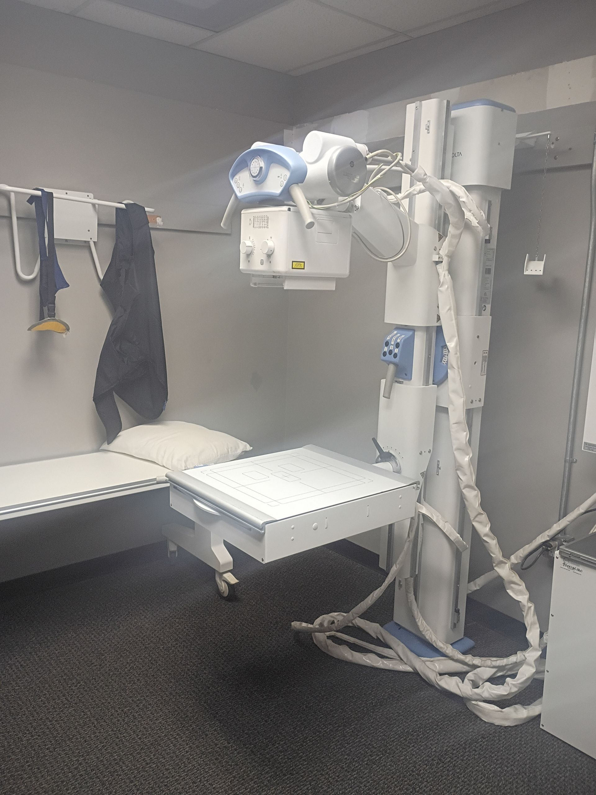 X-ray machine in a medical room.  A table and equipment are visible. Dark carpet, white walls.