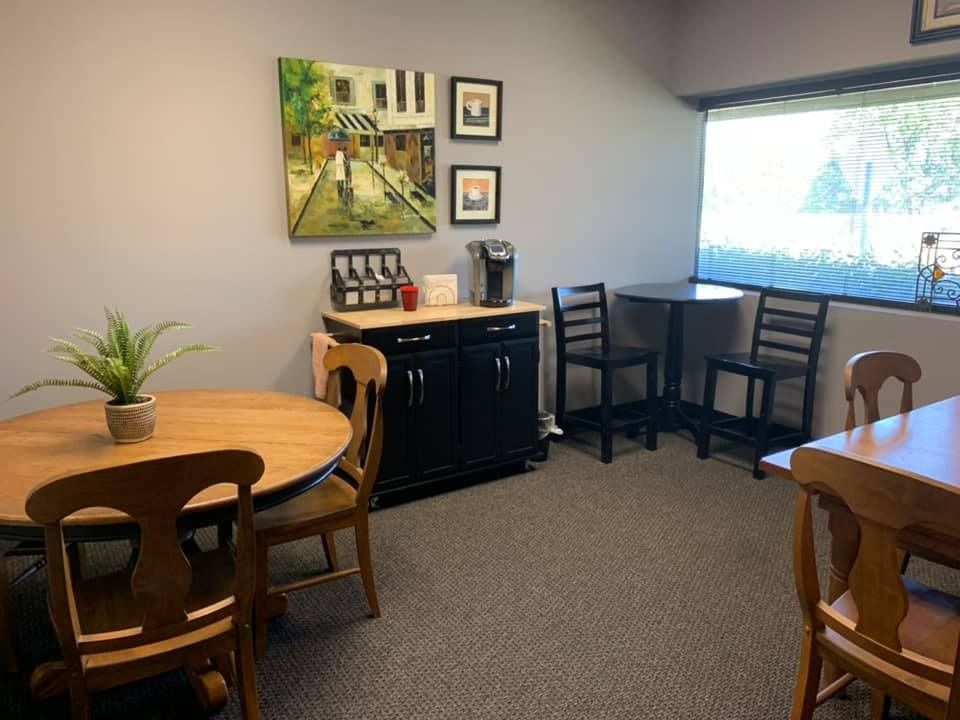 Cozy office break room with coffee station, small tables, chairs, and artwork. Window provides natural light.
