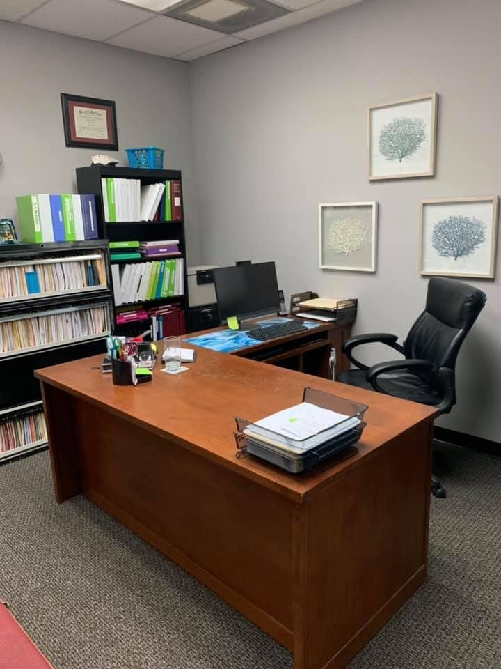 Office with a large wooden desk, black office chair, and bookshelves. Gray walls with framed artwork.
