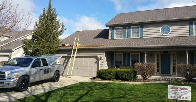 A silver pickup truck parked in a suburban driveway next to a house with a ladder leaning against the roof for repairs.
