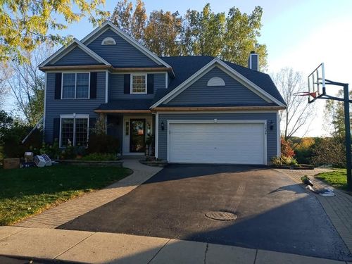 A two-story, blue suburban house with a white garage door, concrete driveway, and a basketball hoop on the right.