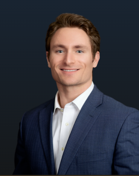 Man in a navy blue suit and white shirt, smiling, against a dark blue background.