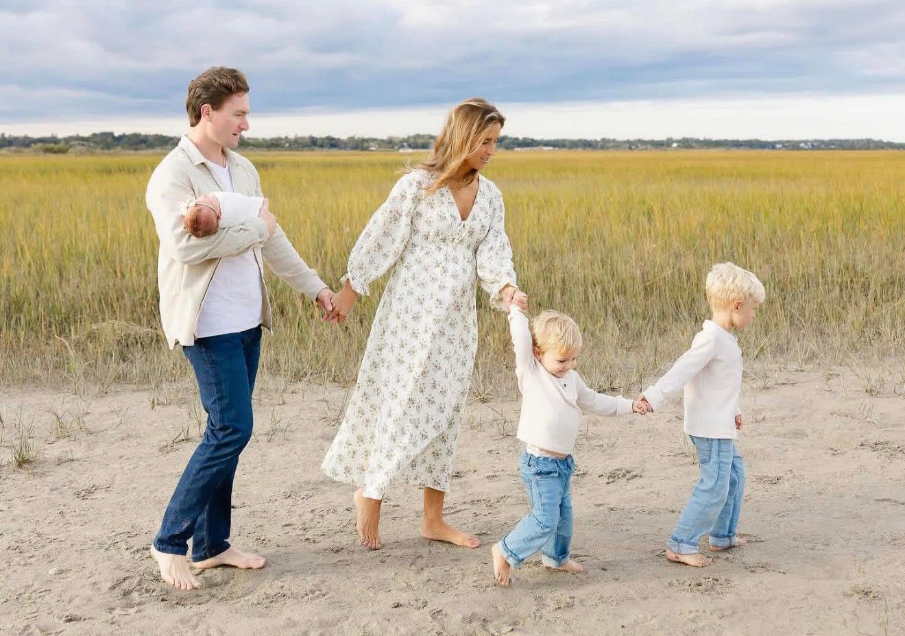 Family walking on sandy path, holding hands; grassy field in background.