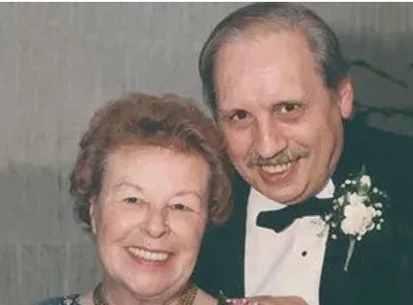 Smiling couple; woman with auburn hair and man in a tuxedo, posing for a formal photo.