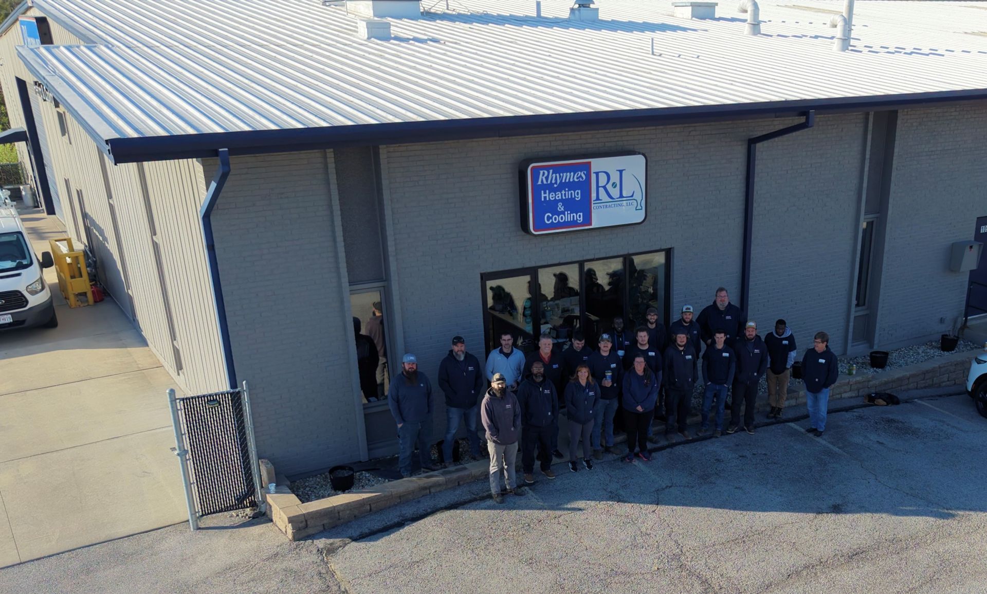 Group of people gathered outside a building entrance under a blue sign, standing on a gravel lot.