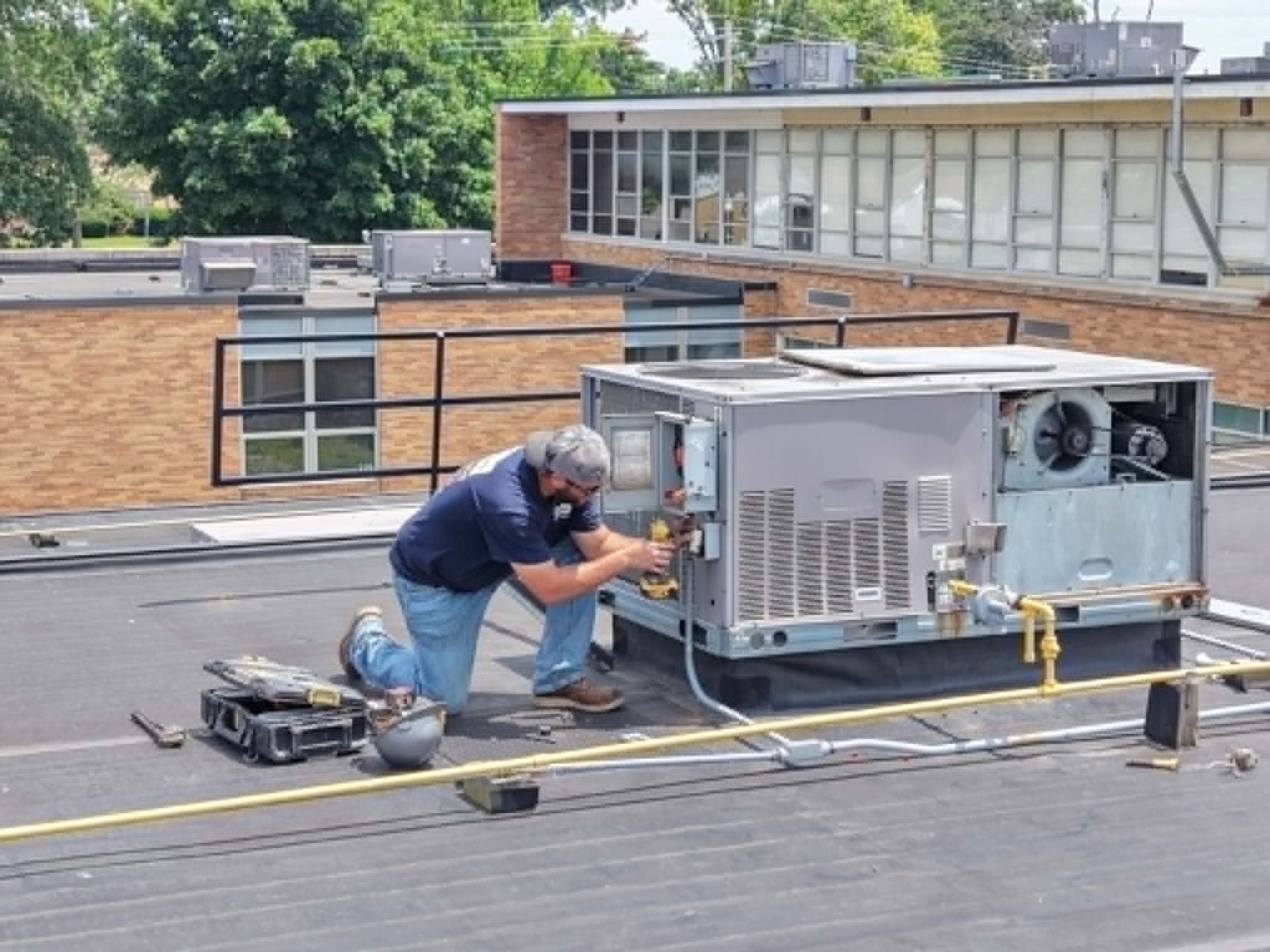 Technician kneeling on a rooftop, repairing an HVAC unit beside tools and safety lines.