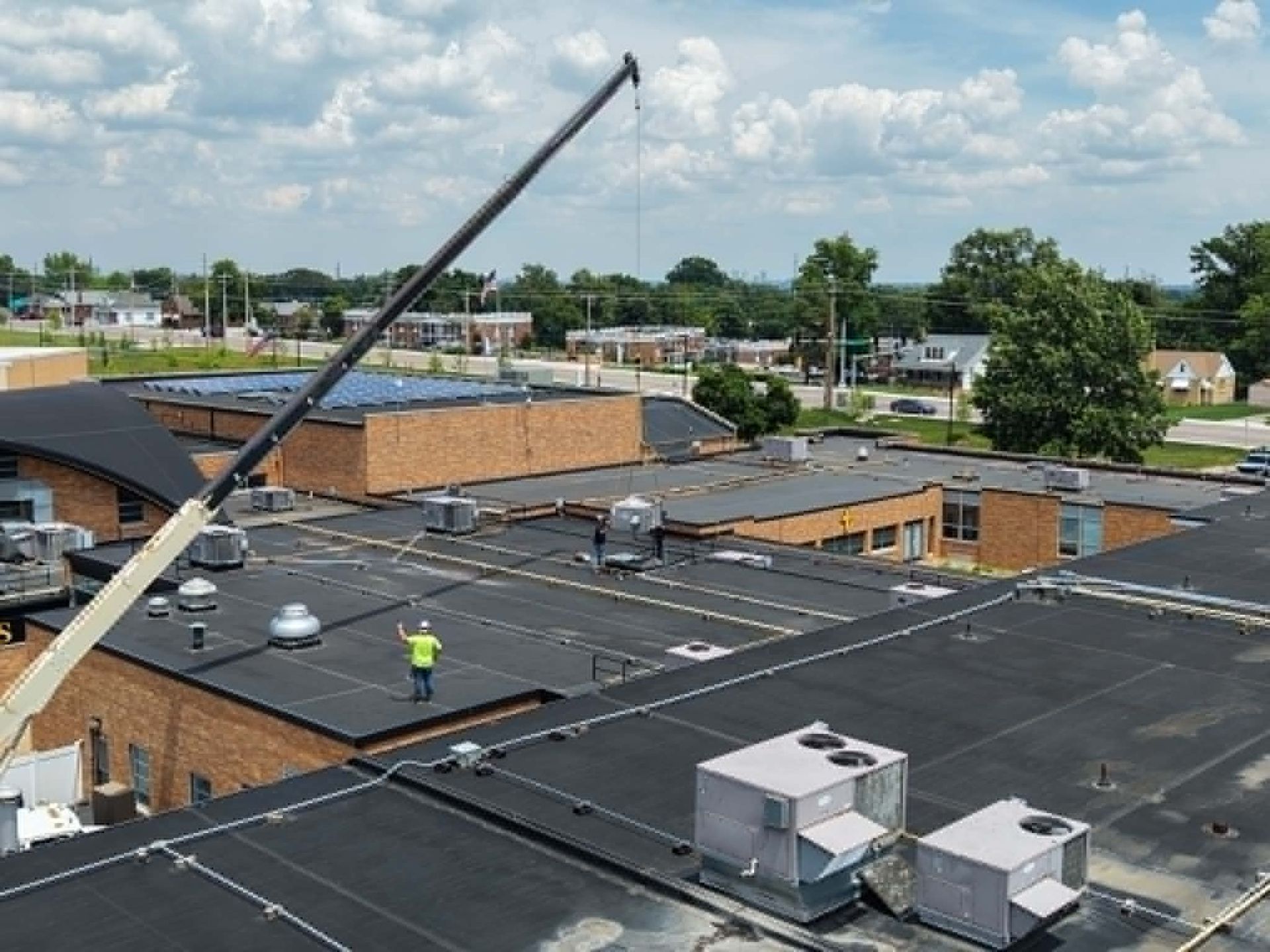 Rooftop with workers, crane boom, and HVAC units on a flat commercial building under a cloudy sky