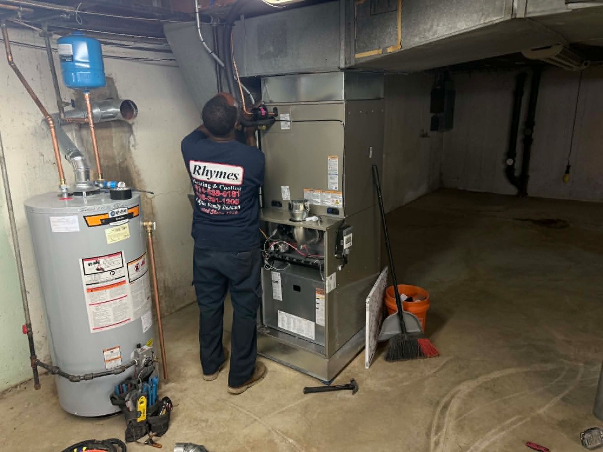 Technician servicing a water heater and furnace in a basement utility room.