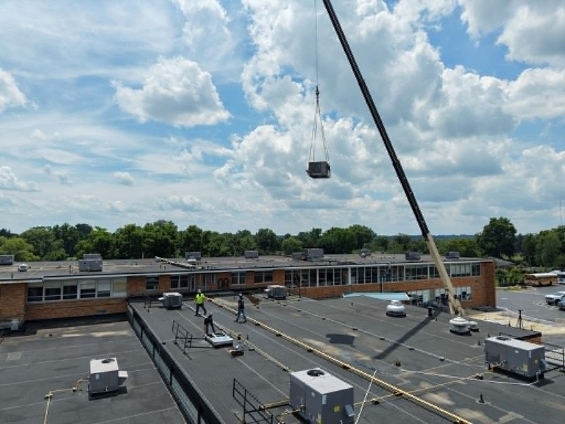 Roof workers and a crane lifting HVAC unit on a flat commercial building under partly cloudy sky