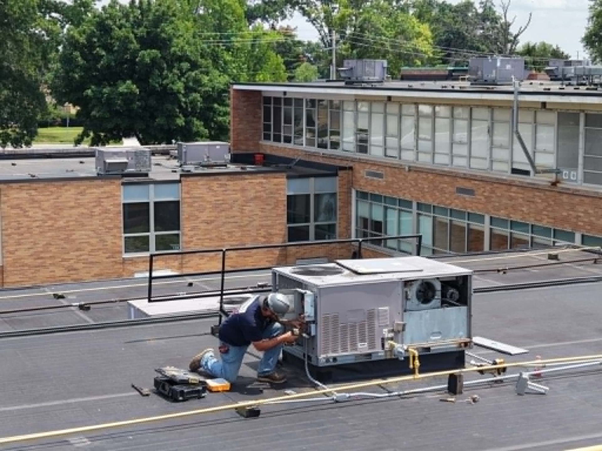 Worker crouches on a rooftop beside HVAC equipment, repairing a unit near a brick building.