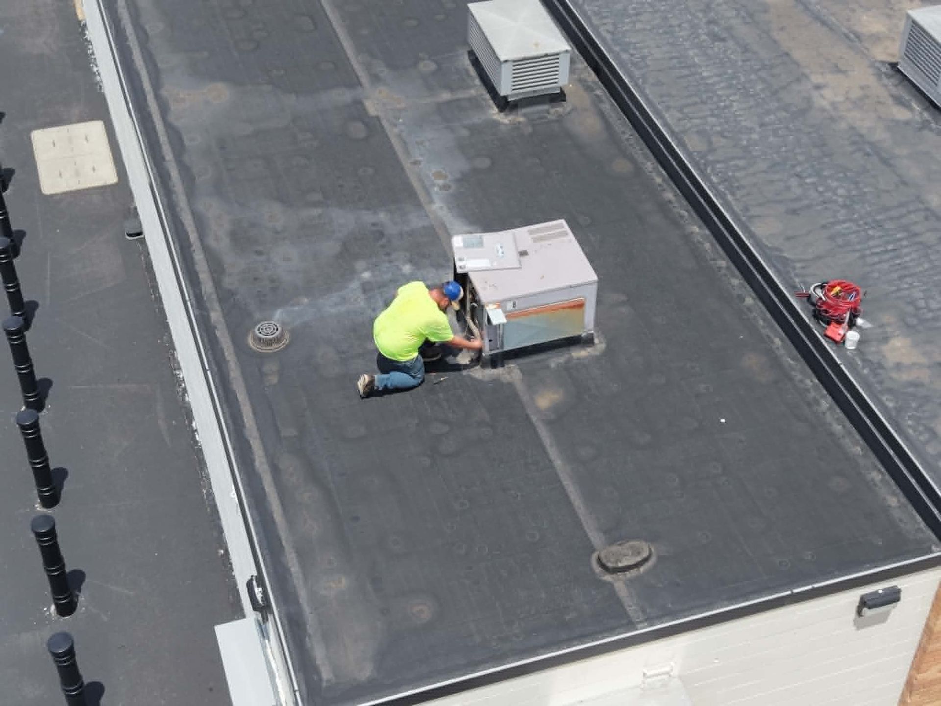 Worker in a neon jacket kneeling on a rooftop beside an HVAC unit