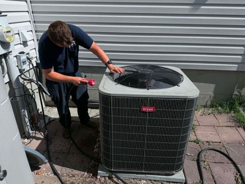 Technician spraying cleaner into an outdoor air conditioner unit beside a house siding wall