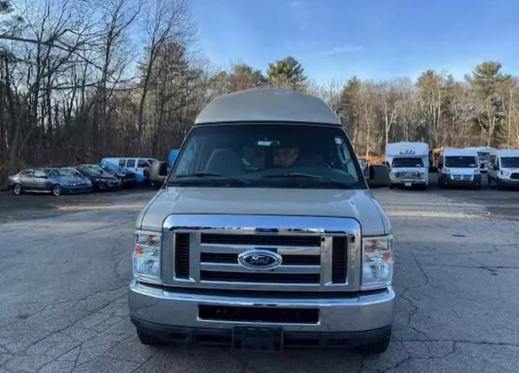 Front view of a beige Ford conversion van parked on an asphalt lot with other vehicles in the background.