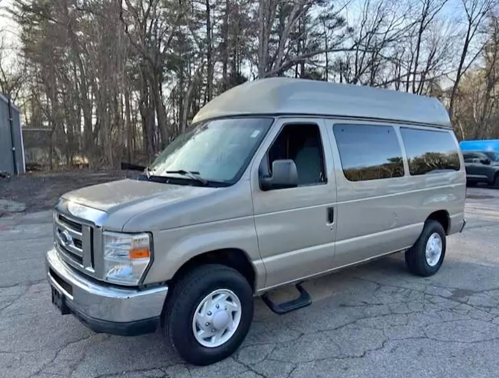 A tan Ford Econoline van with a high roof parked outside on an asphalt lot with trees in the background.