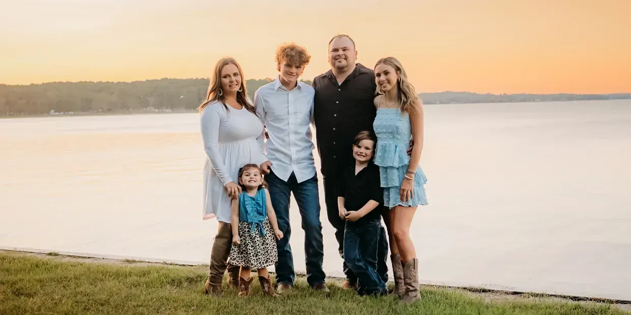 Family of six stands on a grassy bank with water and a sunset in the background.