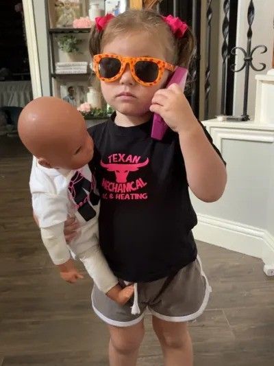 Young girl in sunglasses, holding a baby doll and a phone, wearing a black shirt and shorts.