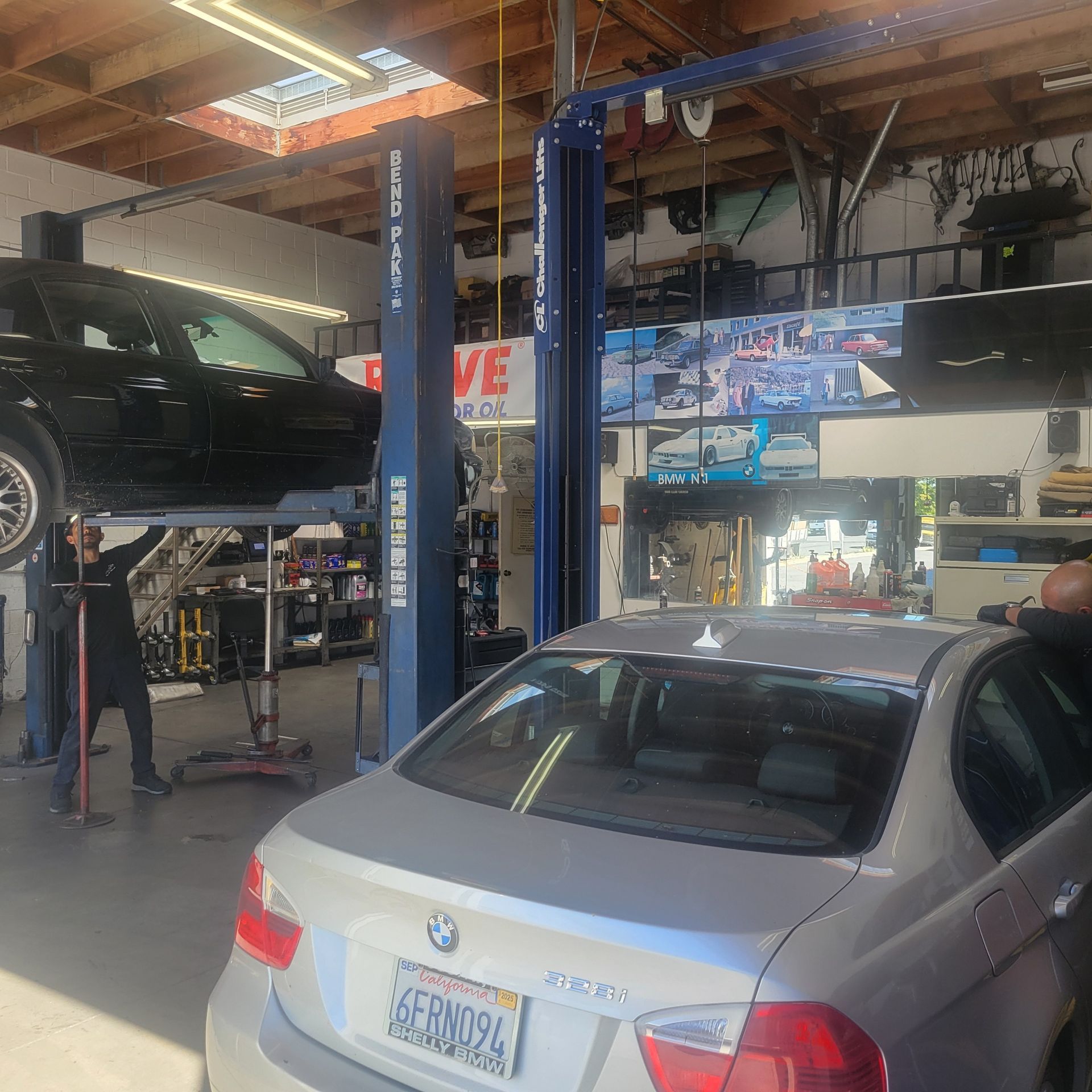 Two mechanics work on a black car lifted on a hydraulic hoist inside a repair shop with a silver BMW parked in the front.
