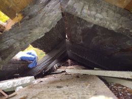 A gloved hand reaches into a charred wooden attic space, revealing burnt rafters and debris from fire damage.