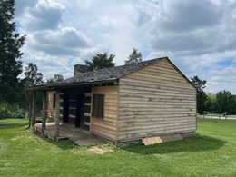 A rustic log cabin with a stone chimney stands in a grassy field under a sunny sky.