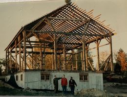 Three people stand in front of a partially constructed wooden barn frame set atop a concrete foundation.