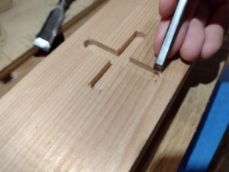 A hand uses a marking knife to outline a cross-shaped mortise on a light-colored wooden board.