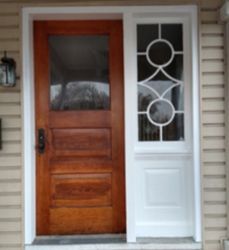 A wooden front door with a glass pane next to a white side panel featuring a decorative geometric window design.