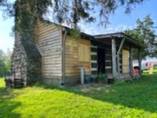 A rustic log cabin with a stone chimney stands in a grassy field under a sunny sky.
