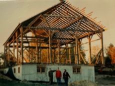 Three people stand in front of a partially constructed wooden barn frame set atop a concrete foundation.