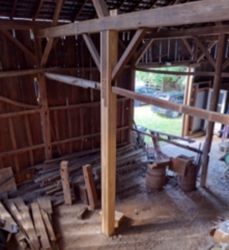 Interior view of a rustic wooden barn with exposed beams and a central support post, looking out toward a grassy yard.